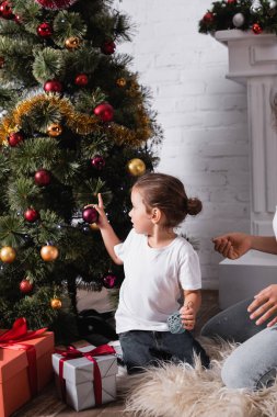 Evde hediye kutularının yanında çam ağacı süsleyen Noel topları olan bir kız.