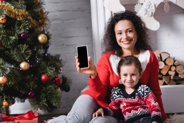 Kız ve anne, süslü çam ağacının yanında boş ekranlı akıllı telefon gösteriyor.