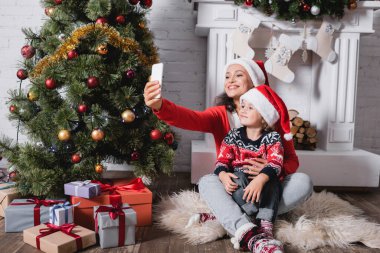 Noel Baba şapkalı anne ve kız evde çam ağacının yanında selfie çekiyorlar.