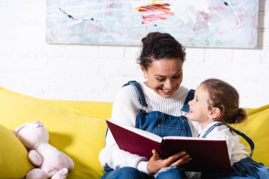 Anne evde sarı koltukta kızının yanında kitap tutuyor.