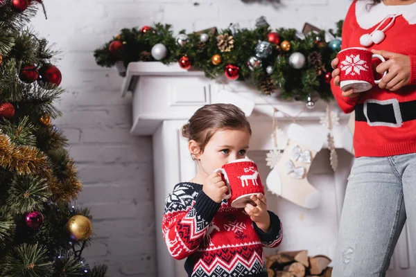 Niña Bebiendo Taza Pie Con Madre Cerca Chimenea Casa — Foto de Stock