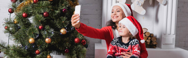 Panoramic shot of mother and daughter in santa hats taking selfie near pine