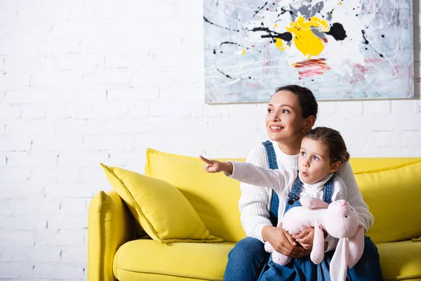 Mãe Abraçando Filha Segurando Brinquedo Macio Apontando Com Dedo Casa — Fotografia de Stock