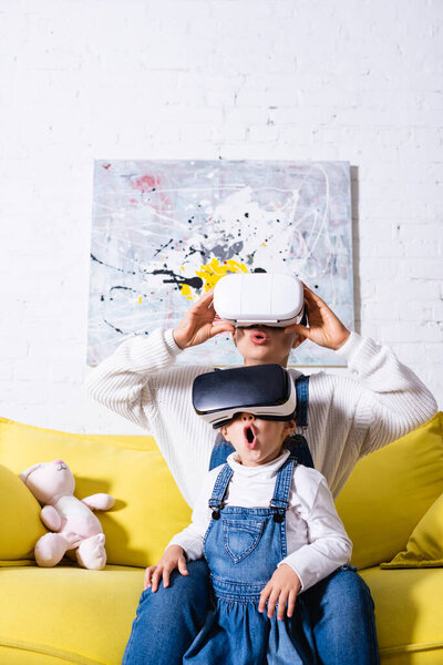 Mother and daughter using virtual reality headsets sitting on yellow couch
