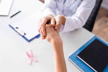 Selective focus of doctor holding patient hand near pink ribbon on table clipart