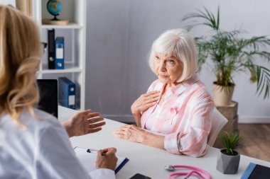 Selective focus of elderly woman with pink ribbon sitting near doctor in clinic clipart