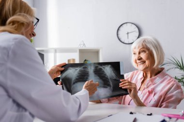 Pembe kurdeleli yaşlı bir kadın ve iş yerinde göğüs röntgeni çeken bir doktor.