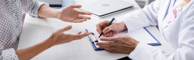 Panoramic shot of patient and doctor writing on clipboard at workplace clipart