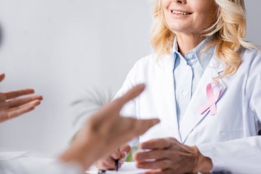 Pembe kurdeleli beyaz önlüklü doktorun seçici odak noktası hastanın yanında oturuyor.