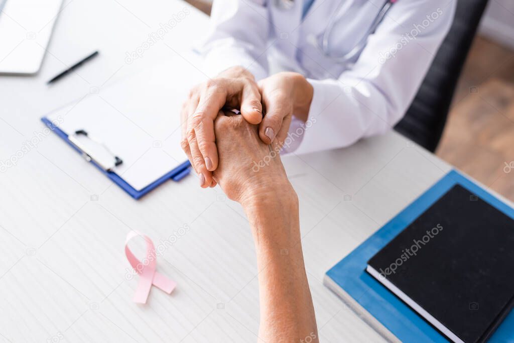 Selective focus of doctor holding patient hand near pink ribbon on table