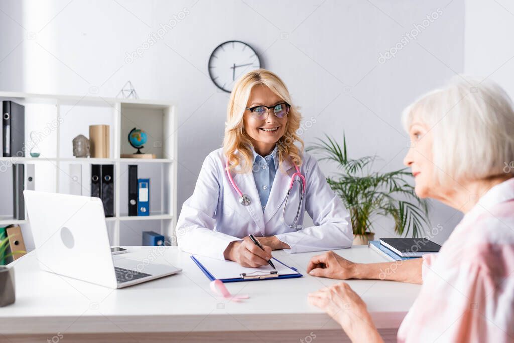 Selective focus of doctor writing on clipboard and looking at patient in clinic