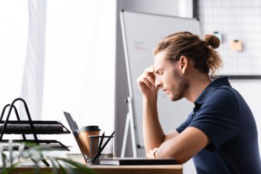 Gözü kapalı düşünceli bir ofis çalışanı laptopun yanındaki çalışma masasında arka planda bulanık bitkilerle oturuyor.