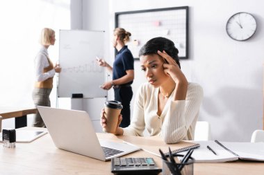 Kağıt bardaklı yorgun Afrikalı Amerikalı kadın iş yerinde bilgisayarına bakıyor ve arka planda bulanık ofis çalışanları var.