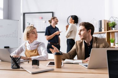 Sarışın bir kadınla konuşurken, arka planda gülümseyen, çok kültürlü ofis çalışanlarıyla el kol hareketi yapan bir adam.