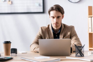 Ofis çalışanı laptopa bakarken ofisteki kırtasiyede bulanık arka planda masasında oturuyor.