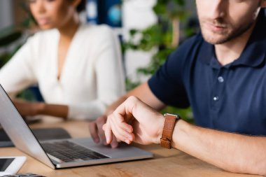 Ofiste bilgisayarla arka planda bulanık bir Afrikalı Amerikalı kadın varken saate bakan ofis çalışanının görüntüsü kesilmiş.