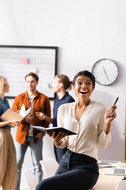 Heyecanlı Afrikalı Amerikalı iş kadını elinde defter ve kalemle çok kültürlü meslektaşlarının yanında bulanık arka plan hakkında konuşuyor.