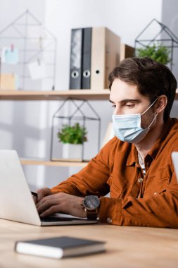 Tıbbi maskeli genç işadamı bulanık önplanda dizüstü bilgisayarda daktilo kullanıyor.