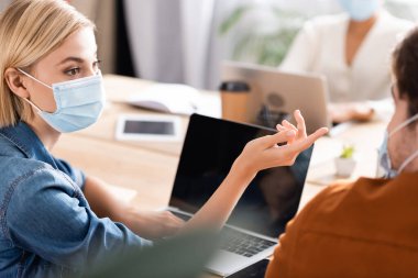 Tıp maskeli bir iş kadını meslektaşı ve laptopunu işaret ediyor. Önplanda bulanıklık var.