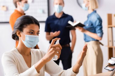 african american businesswoman spraying antiseptic on hands near colleagues on blurred background clipart