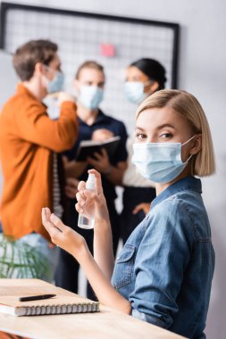 Tıbbi maskeli sarışın müdür elinde antiseptik tutarken meslektaşları arka planda bulanık konuşuyor.