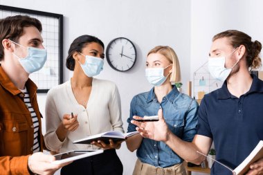 Tıp maskeli çok ırklı iş adamları ellerinde defterler ve ofiste konuşuyorlar.