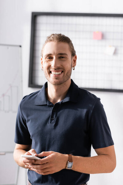 Happy office worker with smartphone laughing, while looking at camera at workplace on blurred background