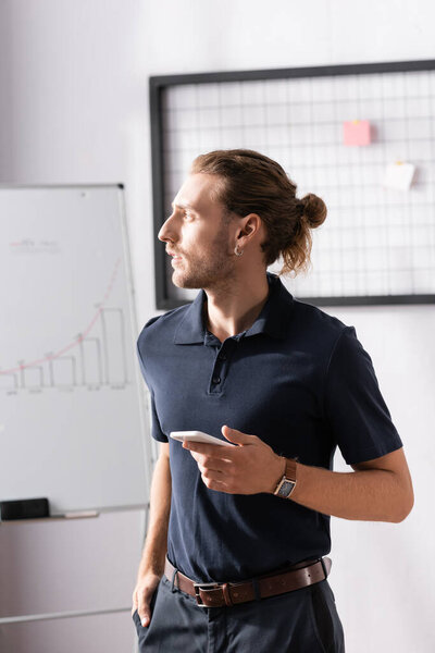 Confident businessman with smartphone looking away while standing in office on blurred background