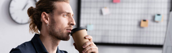Confident young adult man holding paper cup and looking away with blurred mesh organizer on background, banner