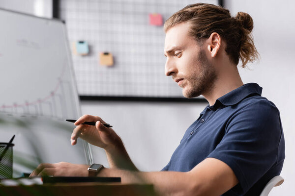 Thoughtful office worker with pen looking away while sitting at workplace desk with blurred plant on foreground