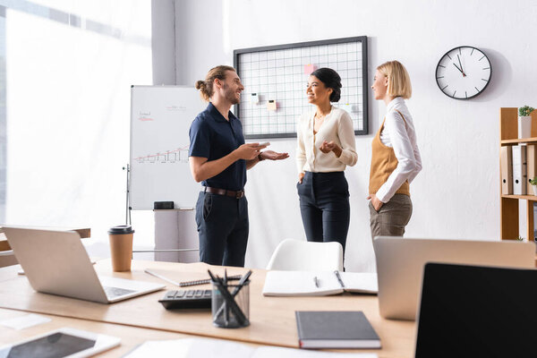 Smiling multicultural female office workers with hands in pocket talking with man while standing near workplace