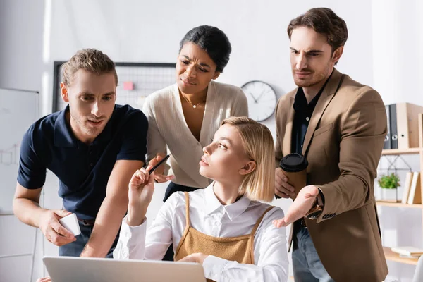 Sarışın kadın şüpheli, çok kültürlü ofis çalışanlarıyla masaya oturmuş iş yerinde dizüstü bilgisayara bakarken başka tarafa bakıyor.
