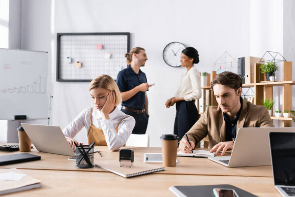 Bored office workers using laptop and writing on notebook while sitting at table, with multicultural colleagues talking on background