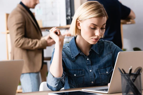 Szőke Tollal Használja Nézi Laptop Miközben Munkahelyen Homályos Férfiak Háttérben — Stock Fotó