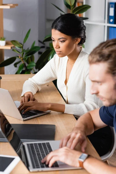 İşyerinde ön planda bulanık bir adamla otururken dizüstü bilgisayara bakan odaklanmış Afrikalı Amerikalı kadın.