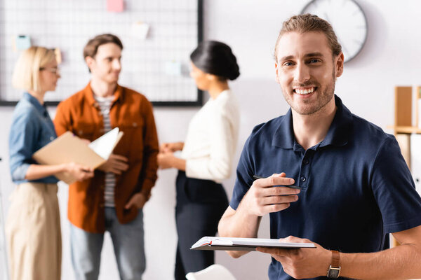 happy businessman looking at camera while multicultural colleagues talking on blurred background