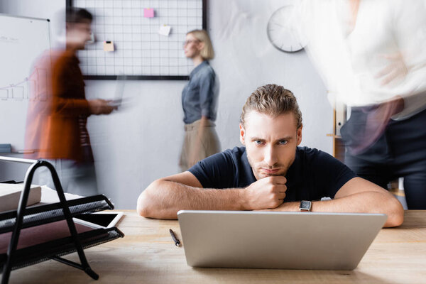 serious manager sitting with head on hands near laptop in open space office, motion blur
