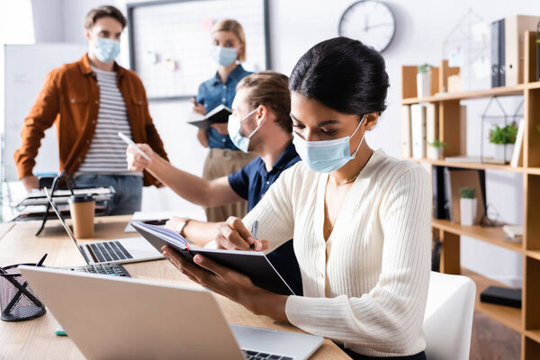 african american businesswoman in medical mask writing in notebook near colleagues working on blurred background