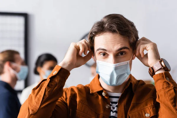Young Manager Putting Medical Mask While Looking Camera Blurred Multicultural — Stock Photo, Image
