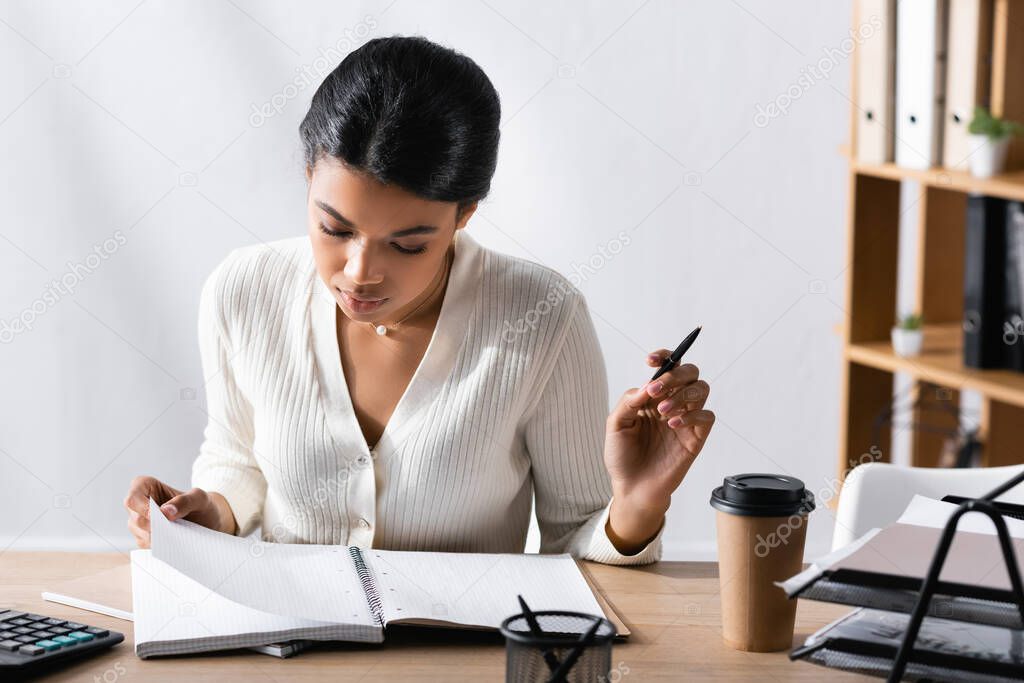Focused african american woman with pen, looking at blank notebook while sitting at desk in office on blurred background