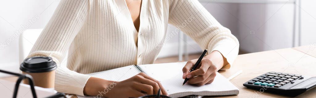 Cropped view of african american woman writing on blank notebook while sitting at desk on blurred background, banner