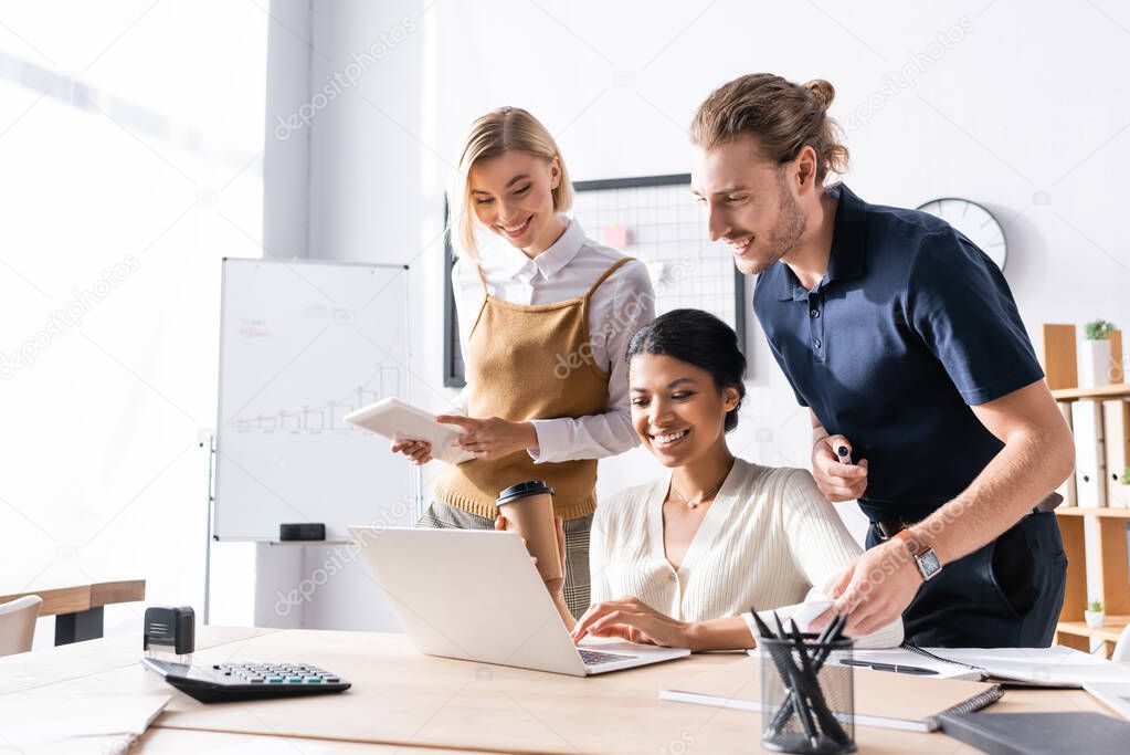 Smiling office workers looking at laptop, while standing near african american woman sitting at table