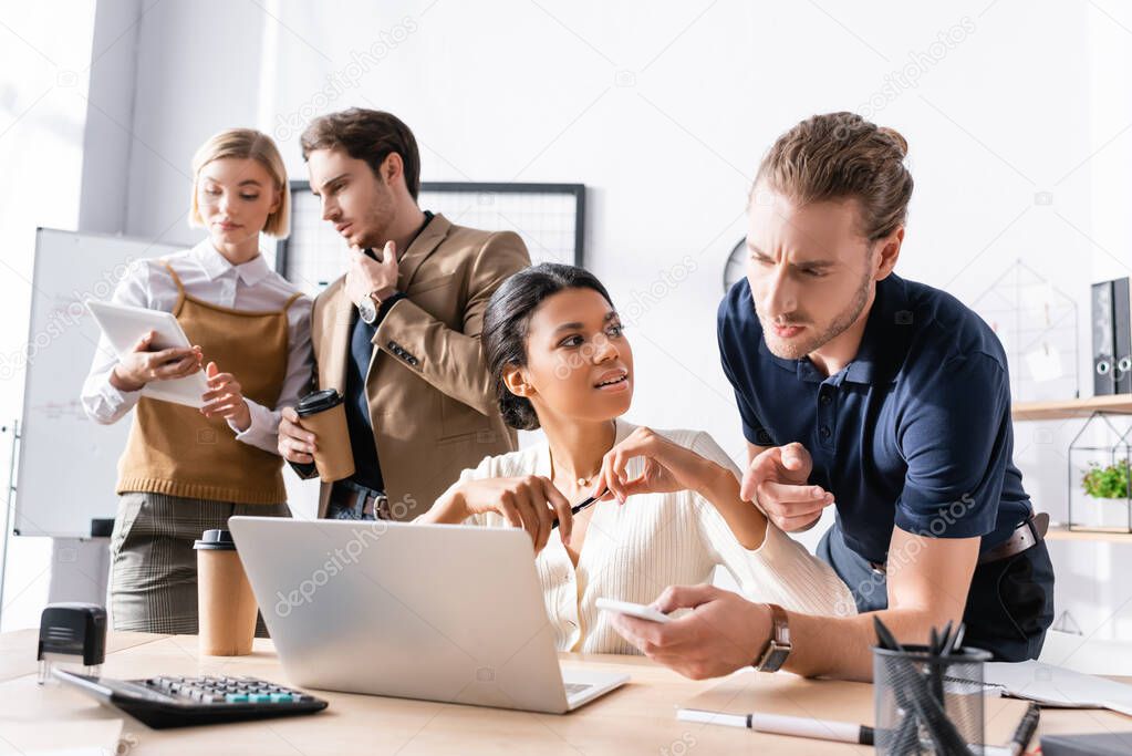 Thoughtful multicultural office workers talking near laptop on table while colleagues working on background in office