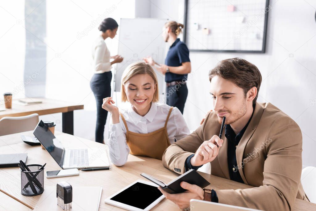 Happy office workers looking at notebook while sitting at table in office with blurred multicultural colleagues working on background