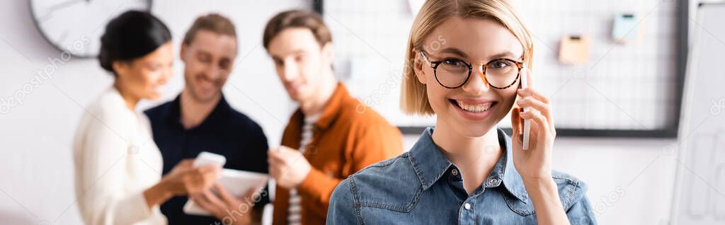 smiling, blonde manager talking on smartphone near multicultural businesspeople on blurred background, banner