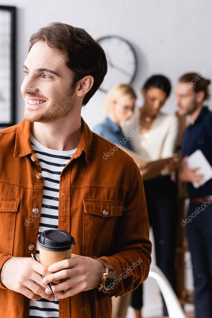 Cheerful manager holding coffee to go and looking away while multiethnic businesspeople talking on blurred background