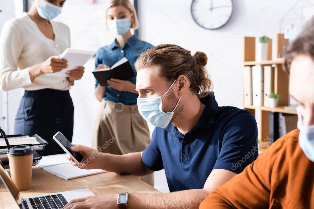 manager in medical mask using smartphone near multiethnic colleagues working on blurred background