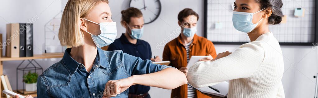 young businesswomen in medical masks doing elbow bump near colleagues on blurred background, banner