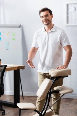 Front view of cheerful masseur standing and pointing with hand at massage chair in office clipart