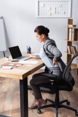 Tired businesswoman with pain in back holding hand on hip, while sitting on office chair in office clipart